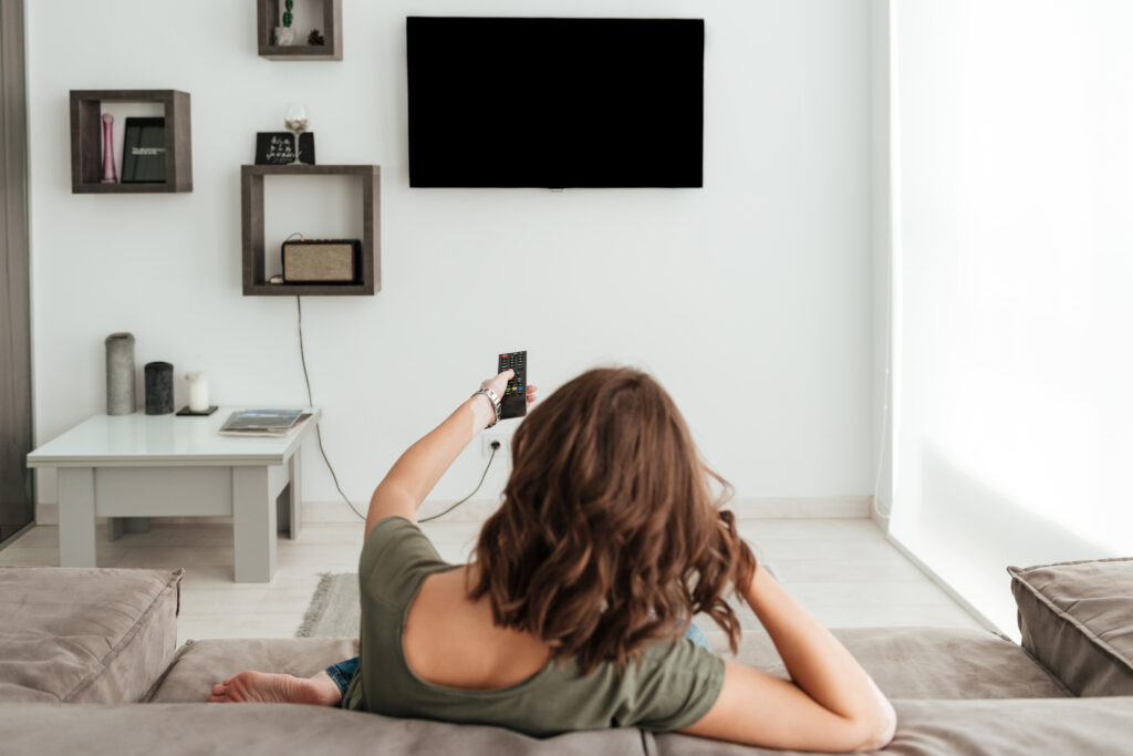 femme assise de dos avec une télécommande dans la main qui allume sa télé